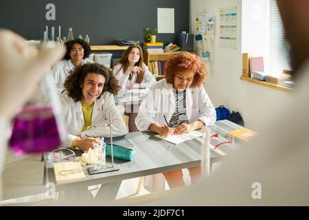 Glücklicher Teenager im Laborkittel, der den Lehrer für Chemie ansieht, der während des Experiments eine Röhre mit einer Mischung chemischer Substanzen zeigt Stockfoto