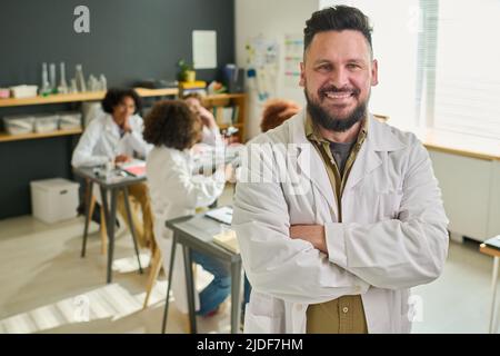 Glücklicher bärtiger, reifer Chemielehrer im Labcoat, der seine Arme auf der Brust kreuzte und die Kamera gegen eine Gruppe von Teenagern ansah Stockfoto