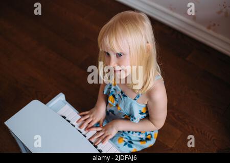 Kleines blondes Mädchen spielt zu Hause ein Spielzeug-Klavier. Musikalische Entwicklung in der Kindheit Stockfoto