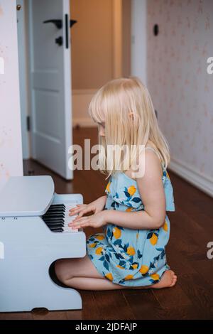 Kleines blondes Mädchen spielt zu Hause ein Spielzeug-Klavier. Musikalische Entwicklung in der Kindheit Stockfoto