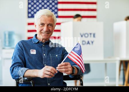 Vorderansicht Porträt eines lächelnden älteren Mannes mit Behinderung, der die US-Flagge in der Wahlstation hält, Kopierraum Stockfoto