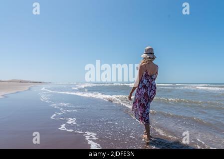 Frau in Sommerkleidung, die barfuß an einem windigen Strand entlang schlendert und mit einem Stockfoto