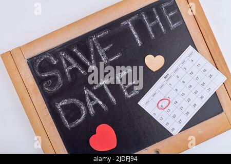 Speichern Sie die Datumswörter auf der Tafel. Kalender mit eingekreistem Datum und Papierherzen. Stockfoto