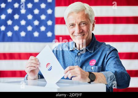 Porträt eines lächelnden älteren Mannes, der gegen die amerikanische Flagge den Stimmzettel in den Papierkorb legt, Platz zum Kopieren Stockfoto