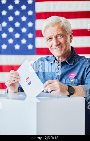 Vertikales Porträt eines lächelnden älteren Mannes, der im Hintergrund gegen die amerikanische Flagge den Stimmzettel in den Papierkorb legt Stockfoto