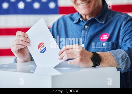 Nahaufnahme eines lächelnden älteren Mannes, der im Hintergrund gegen die amerikanische Flagge den Stimmzettel in den Papierkorb legt, Platz zum Kopieren Stockfoto