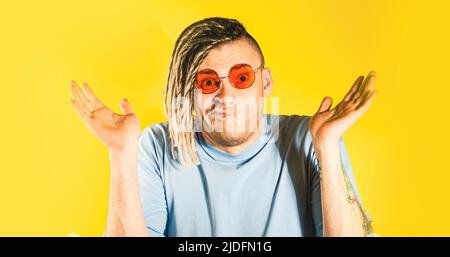 Junger, gutaussehender Mann mit Sonnenbrille, blauem T-Shirt mit einer Geste des Nichtwissens und Blick auf die Kamera. Ratlos Kerl spreizte Hände auf gelbem Hintergrund i Stockfoto