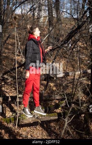 Große Frau in rotem Sportanzug, die auf Baumstämmchen über die Grube geht und im Wald weg von der Zivilisation, näher an der Natur, mit dem sie lächelnd davonschaut Stockfoto