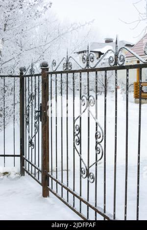 Schmiedeeiserner gemusterter verzierter Zaun mit Tür, die mit leichter Schneeschicht bedeckt ist, in ein privates Gebiet mit Schnee auf dem Boden und Ästen von Bäumen Stockfoto