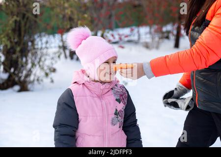 Nahaufnahme der weiblichen erwachsenen Hand, die Karotte auf das Gesicht der Tochter als Nase auf dem Hinterhof voller Schnee legte, nachdem sie abends mit Bäumen und Eisenzaun spazieren ging Stockfoto