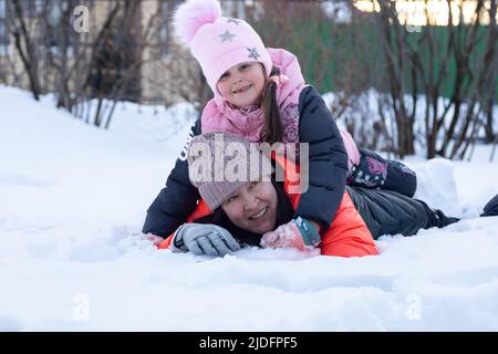 Kaukasische glückliche kleine Tochter mit Mutter, die auf Schnee liegt, lächelnd im Park am Abend mit Bäumen im Hintergrund. Eltern verbringen Zeit mit Stockfoto