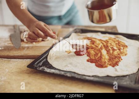 Eine Frauenhand gießt Tomatensauce über die Oberfläche einer Pizza, die auf dem Backblech liegt Stockfoto