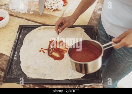 Eine Frau verteilt Tomatensauce auf Pizzateig auf einer Backform Stockfoto