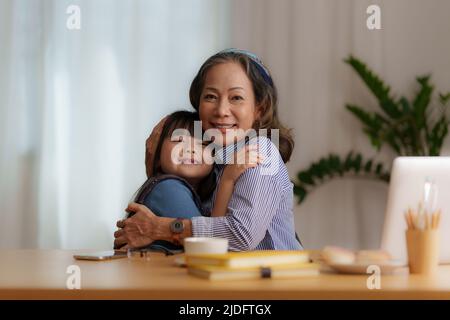 Glückliche Momente der Enkelin umarmen sich, während eine asiatische Geschäftsfrau zu Hause arbeitet. Familienkonzept. Stockfoto