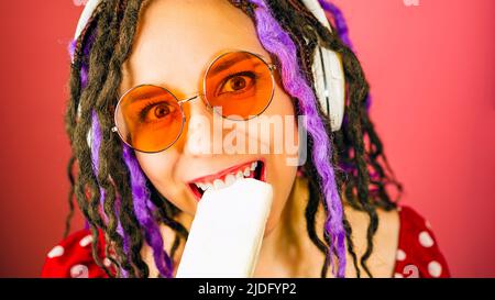 Junge Frau in Sonnenbrillen, Kopfhörer beißen Eis, lächelnd, Blick auf die Kamera. Fröhliche Hündin mit Dreadlocks, die Eisbecher fressen, Musik hören Stockfoto