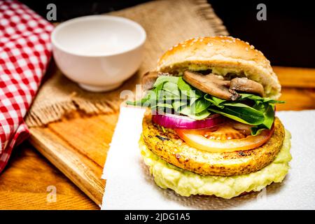 Veganer Veggie-Protein-Burger mit Tomaten, Rucola, roten Zwiebeln, Pilzen und Avocado-Mayonnaise auf einem Holzbrett und schwarzem Hintergrund. Gehackte Ansicht. Stockfoto