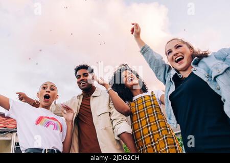 Blick aus der unteren Perspektive auf Freunde, die auf dem Dach feiern. Gruppe von glücklichen jungen Menschen jubeln, während sie unter herabfallenden Konfetti stehen. Multikulturelle Freunde hav Stockfoto