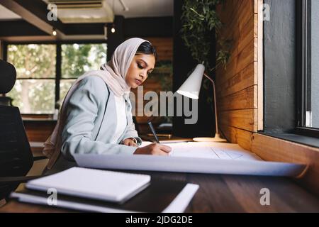 Kreativer muslimischer Designer, der in einem modernen Büro mit Architekturzeichnungen arbeitet. Junge Geschäftsfrau plant ein innovatives Projekt. Architektin Stockfoto