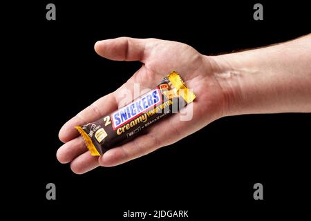 Ein Mann bietet eine Schokoriegel von Snickers an, die Hand hält eine Schokoriegel mit schwarzem Hintergrund in Nahaufnahme. Stockfoto