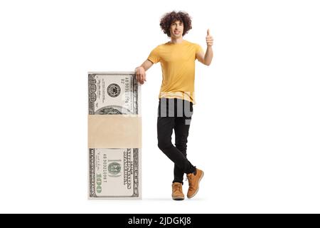 Junger Mann, der sich auf einen Geldstapel stützt und auf weißem Hintergrund die Daumen hoch zeigt Stockfoto