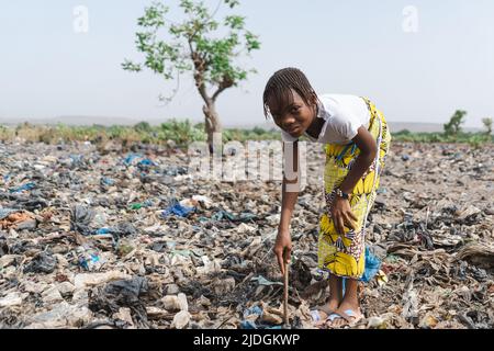 Unschuldiges kleines afrikanisches Mädchen, das recycelbare Abfälle auf einer Deponie sammelt und lächelt, ohne sich der Ausbeutung bewusst zu sein; Kinderarbeit Konzept Stockfoto