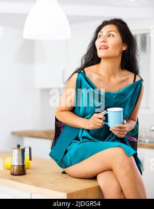 Porträt einer Frau, die in der Küche auf einem Tisch sitzt und Kaffee trinkt Stockfoto