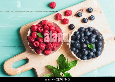 Draufsicht Frische Blaubeeren und Himbeeren in Schüsseln auf einem Schneidebrett auf blauem Holztisch. Gesund essen, veganes Essen flach legen. Stockfoto