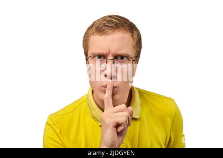 Lustiger junger Mann in gelbem T-Shirt, der um Ruhe bittet, schweigende Geste isoliert auf weißem Hintergrund. Junger Mann mit Brille, der Shhh sagt: Ruhe, bitte Stockfoto