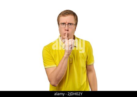 Lustiger junger Mann in gelbem T-Shirt, der um Ruhe bittet, schweigende Geste isoliert auf weißem Hintergrund. Junger Mann mit Brille, der Shhh sagt: Ruhe, bitte Stockfoto