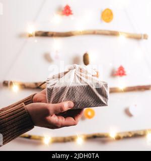 Frauenhände halten die furoshiki Geschenkbox gegen die Dekoration des Weihnachtsbaums aus trockenen Stäbchen, trockener Orange, Zapfen auf weißem Hintergrund. Null Stockfoto