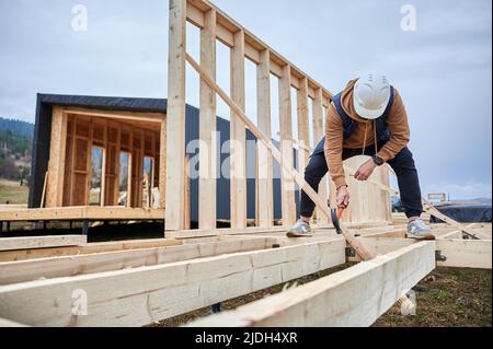 Mann Arbeiter Gebäude Holzrahmenhaus auf Pfahlfundament. Zimmermann hämmert Nagel in Holzbalken, mit Hammer. Schreinerei-Konzept. Stockfoto