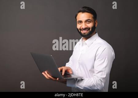 Der indische Unternehmer trägt einen Laptop isoliert auf einem schwarzen, bärtigen Geschäftsmann, der einen Laptop benutzt, tippt, E-Mails empfängt, eine neue Computer-App nutzt, die Kamera anschaut und lächelt Stockfoto