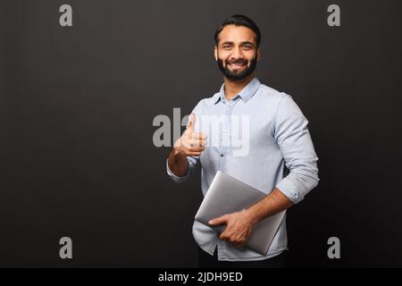 Indischer männlicher Unternehmer im legeren Hemd, der Laptop trägt und den Daumen isoliert auf einem schwarzen, bärtigen Geschäftsmann zeigt, mit Laptop, der auf die Kamera schaut und lächelt, ok Geste Stockfoto