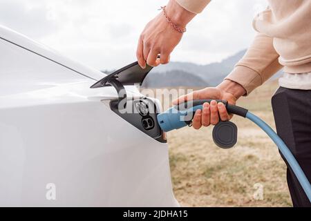 Männliche Hand, die ein Ladegerät in die Steckdose für Elektroautos steckt, im Hintergrund sichtbare Bergnatur Stockfoto