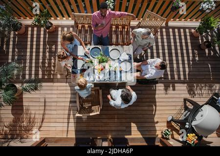 Blick von oben auf die Familie von 3 Generationen, die beim Barbecue-Party-Dinner auf der Terrasse isst, Leute, die am Tisch auf der Terrasse mit Grill sitzen. Stockfoto