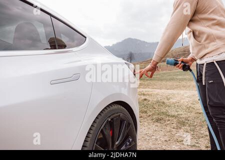 Männliche Hand, die ein Ladegerät in die Steckdose für Elektroautos steckt, im Hintergrund sichtbare Bergnatur Stockfoto