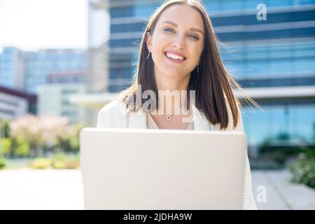 Glückliche attraktive Freiberuflerin oder Studentin mit Laptop im Freien. Fröhliche Frau mit Laptop, die auf die Kamera schaut und lächelt, Büroangestellte mit Laptop Stockfoto