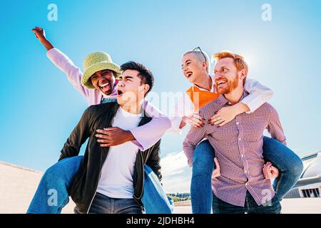 Glückliche junge Männer geben Huckepack Fahrt zu Freundinnen. Gruppe von Freunden genießen einen Tag Stockfoto