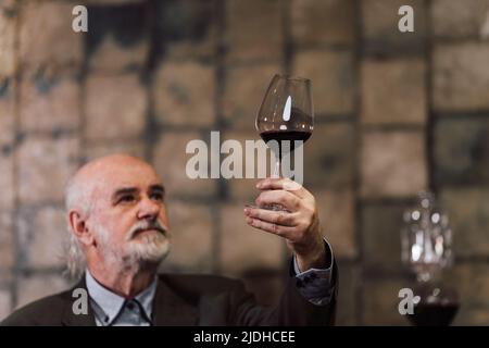 Zufriedener männlicher Kaukasischer Weinexperte, der Rotwein verkostet und sein Glas hochhebt Stockfoto
