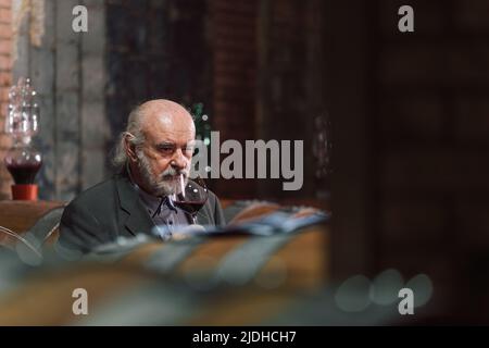 Leitender kaukasischer Mann mit Graubart, ein Weinkenner, der ein Glas hält und den Rotwein durch Anblick, Geruch und Geschmack analysiert Stockfoto