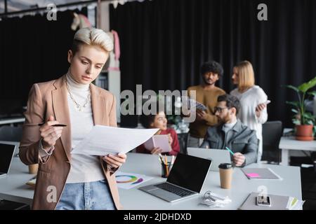 Designer mit Papieren und Stift neben dem Schreibtisch mit Geräten und interrassischen Kollegen auf unscharfem Hintergrund Stockfoto