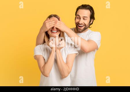 Fröhlicher, froher, tausendjähriger Kaukasianer im weißen T-Shirt schließt die Augen der Frau zur Überraschung Stockfoto