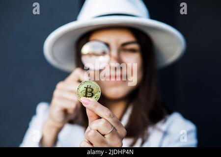 Elegante Geschäftsfrau in einem weißen Hut hält eine Lupe und schaut auf Bitcoin. Stockfoto