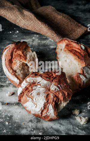 Nahaufnahme eines runden Kamut-Weizenbrots, der in einige Stücke gebrochen wurde, auf einer melierten dunkelgrauen Stoffoberfläche, neben einem Sackleinen Stockfoto