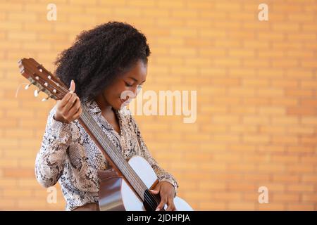 Beschnittene Ansicht einer jungen Frau, die Gitarre spielt und auf einem Backsteinwand-Hintergrund herunterschaut Stockfoto