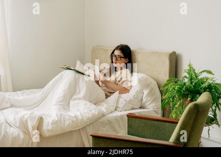 Eine hübsche Studentin, die ein Buch liest und sich Notizen auf dem Handy gemacht hat, während sie im Bett liegt Stockfoto