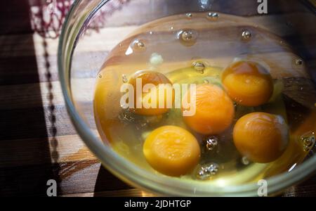 Eier aus der Nähe, Eigelb und Eiweiß in einer Glasschüssel auf einem Bambusbrett. Zubereitung von Rühreiern. Stockfoto