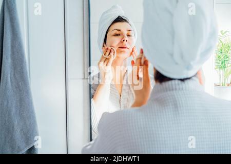 Frau im Bademantel mit einem Handtuch auf dem Kopf Blick in den Spiegel, Peeling Gesichtshaut mit Loofah Schwamm, Entfernen Make-up oder tun Reinigungsroutine, Haut Stockfoto