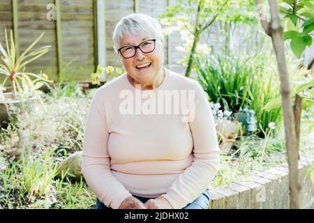 Porträt einer lächelnden älteren, reifen Großmutter im Ruhestand in einer Brille. Glückliche ältere Frau, die lächelt und in ihrem grünen Garten sitzt und den Ruhestand genießt Stockfoto