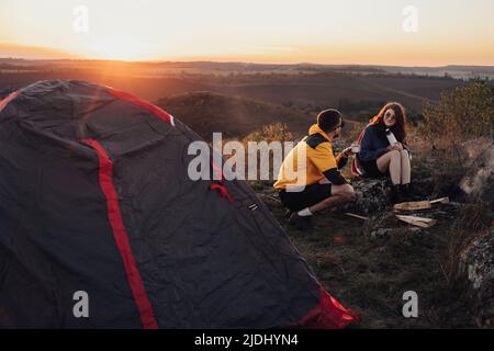 Zwei junge Reisende Mann und Frau sitzen beim Sonnenuntergang über dem Gipfel des Hügels in der Nähe des Lagerfeuers und des Zeltes Stockfoto